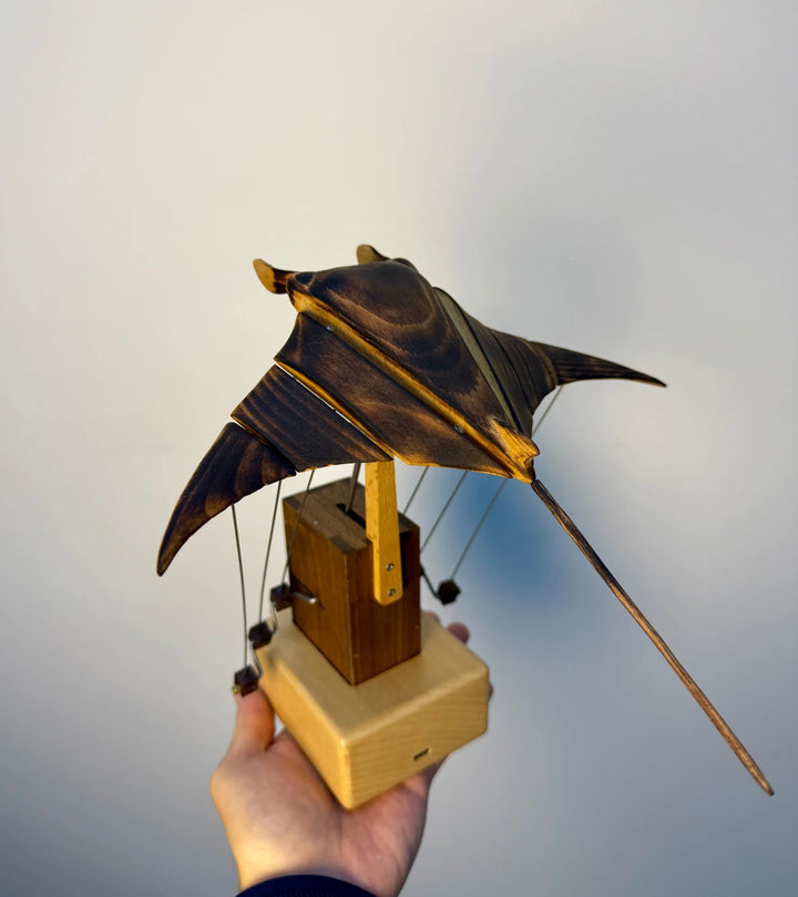 Manta ray automaton held up in one hand for scale, dark tower on a pale yellow wood base.