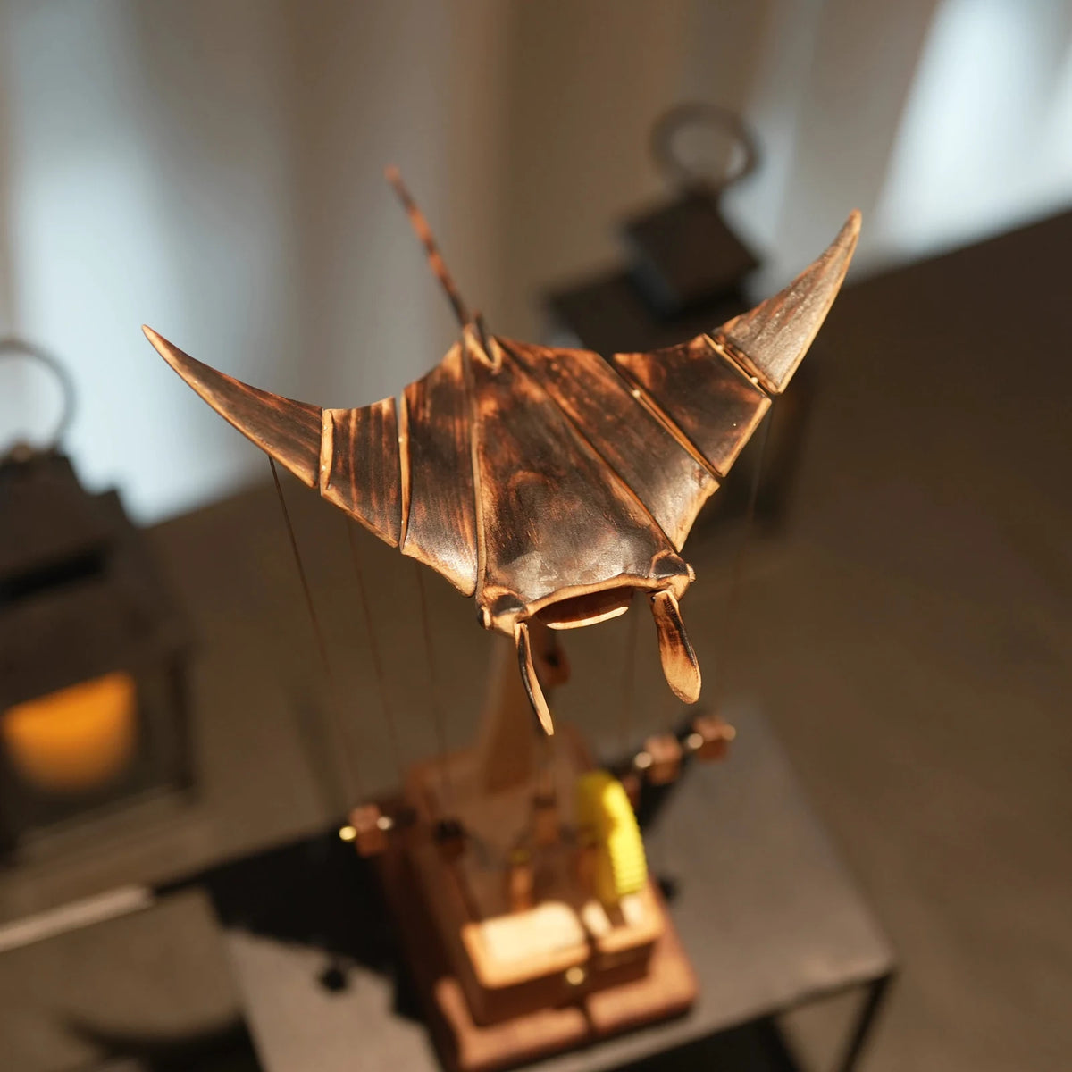 Manta ray automaton on a desk in warm light, a lantern glowing behind and a yellow gear wheel beside its base.