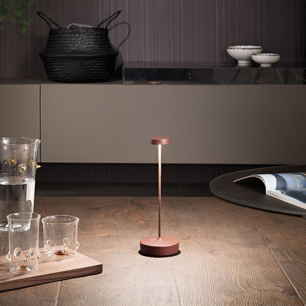 A copper-toned Swap Pro lamp lit on a wooden floor beside glass cups and a magazine