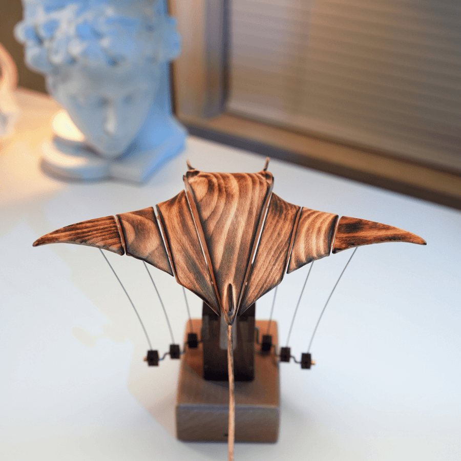 Manta ray automaton from the front with its wings level, a pale sculpted bust blurred behind.