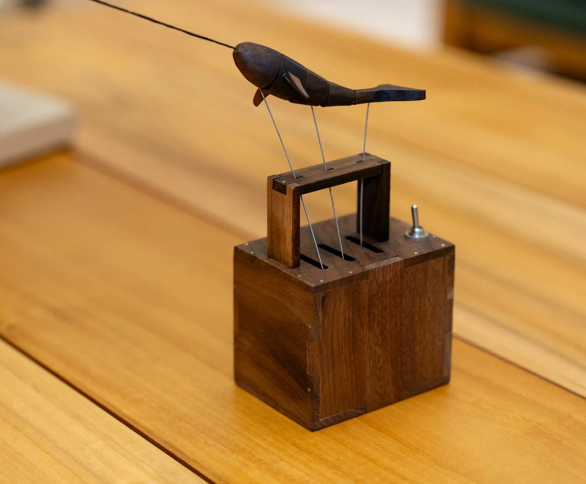 Dark wood narwhal with a long thin horn on fine wires above a small walnut box, on a sunlit wooden table.