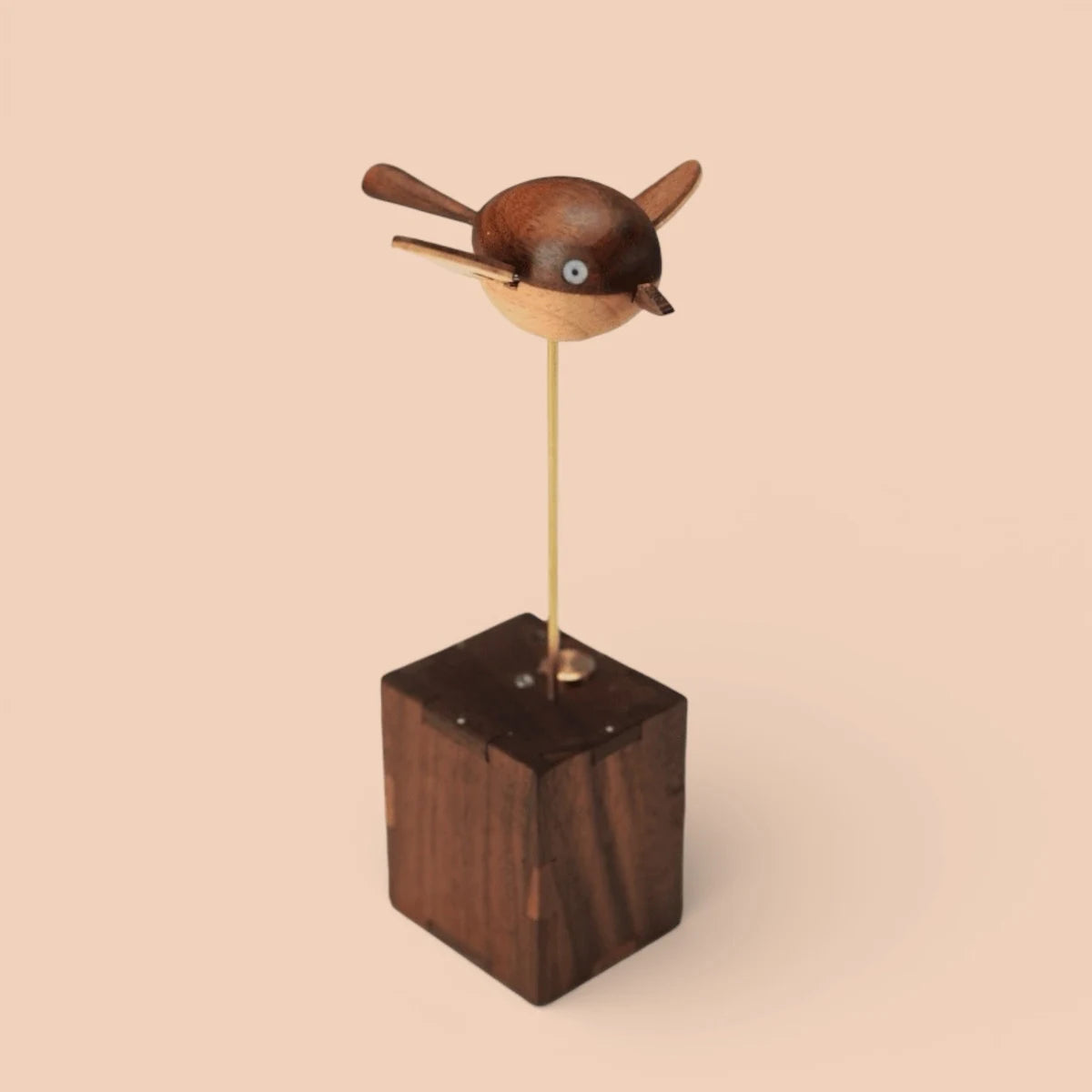 A round dark wood bird with two flat wings and a pale belly on a thin brass rod above a small dark wood box, peach background.