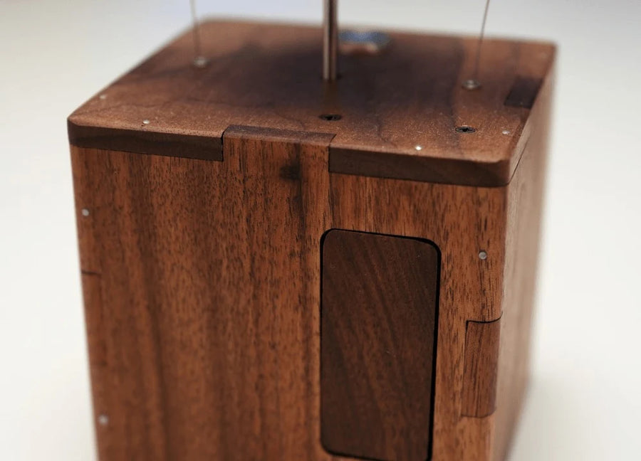 Reddish-brown wood box from the side with a darker recessed rectangular panel on one face, a slim rod and two thin wires rising from the top.