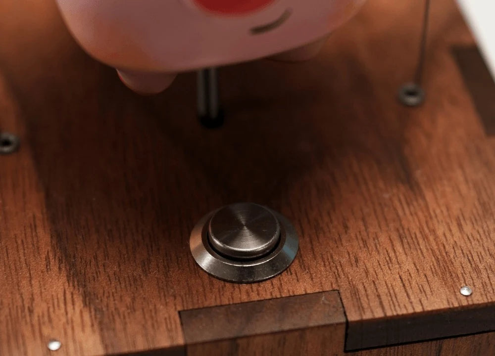 Close view of a round metal button on the wood top of the box, the slim rod rising beside it with the pig's belly above and tiny pins in the wood.