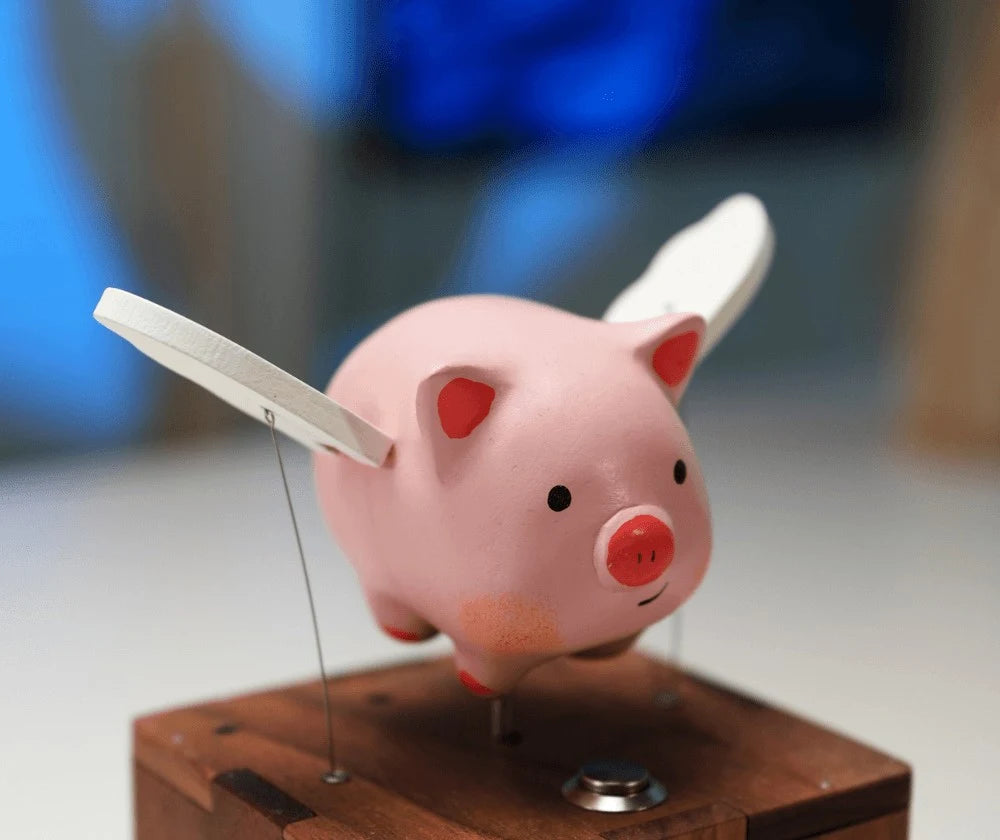 Side view of the pink pig with one white wing swept back and the other pointing forward, thin wires running down to a reddish-brown wood box, blurred blue shapes behind.