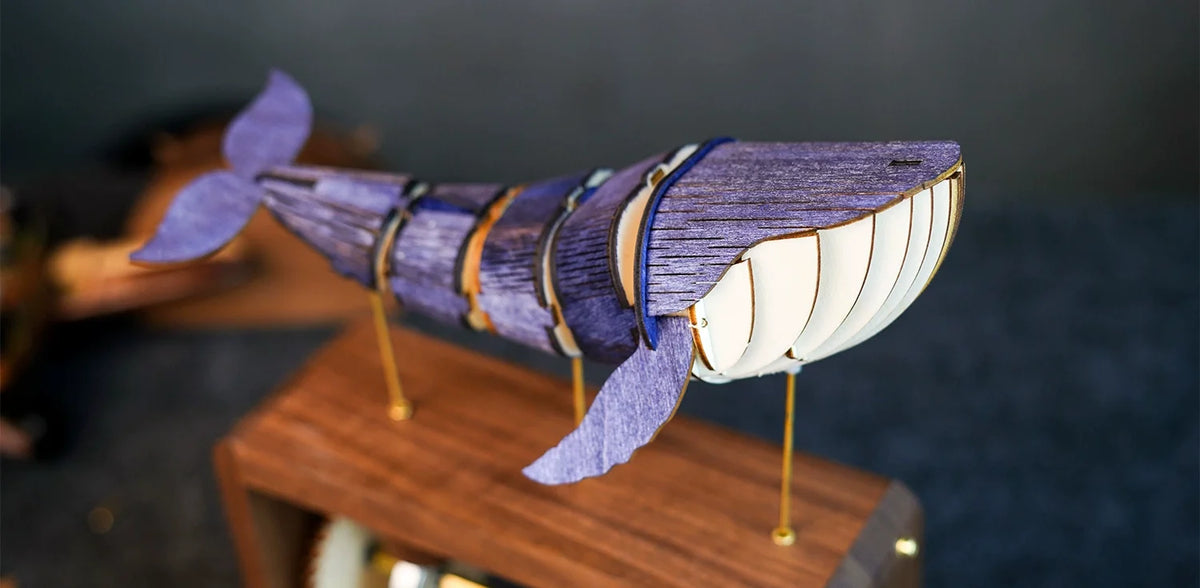 Close side view of a blue-violet wooden whale with pale ribs, standing on brass rods above a walnut box, dark background.