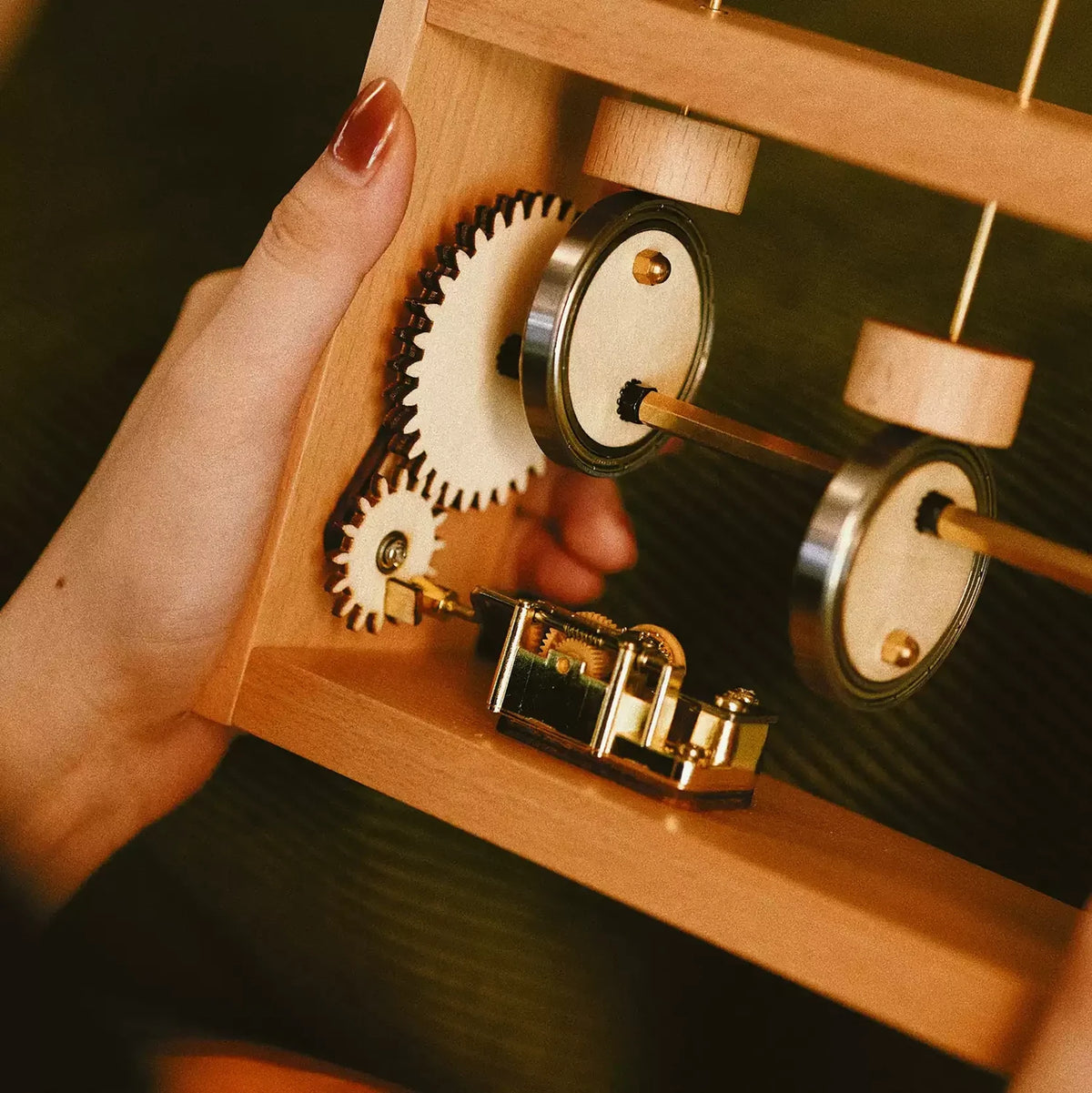 Close view of a beech frame held in one hand: the wooden gear wheels, cams on a brass shaft and the gold music box movement.