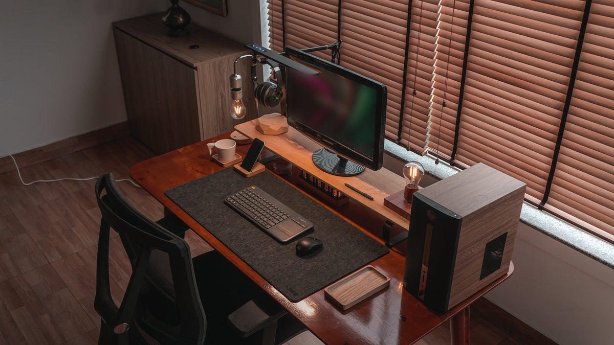 Desk with a monitor on a wooden shelf, floating bulb lamp, wireless keyboard, mouse and a wooden tray beside a window