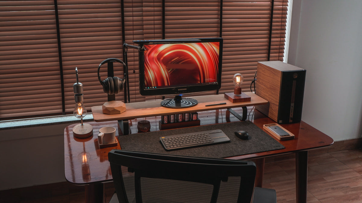 Front view of a desk by a window with a monitor, floating bulb lamps, headphones stand, keyboard and felt desk mat
