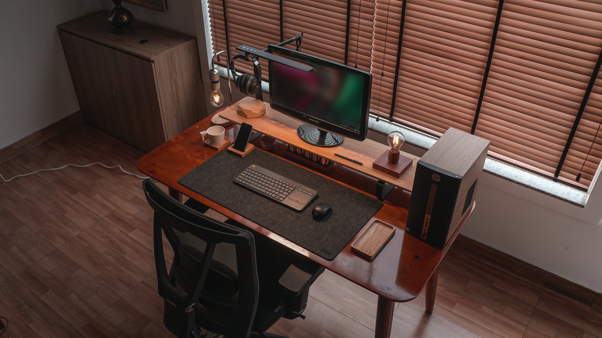 Warm desk setup by a blinds window with a monitor, wooden shelf, floating bulb lamp, keyboard and dark felt mat