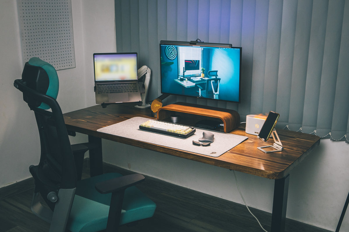 Dark desk setup with a monitor on a wood riser, laptop on an arm, felt mat, keyboard, mouse and phone stand