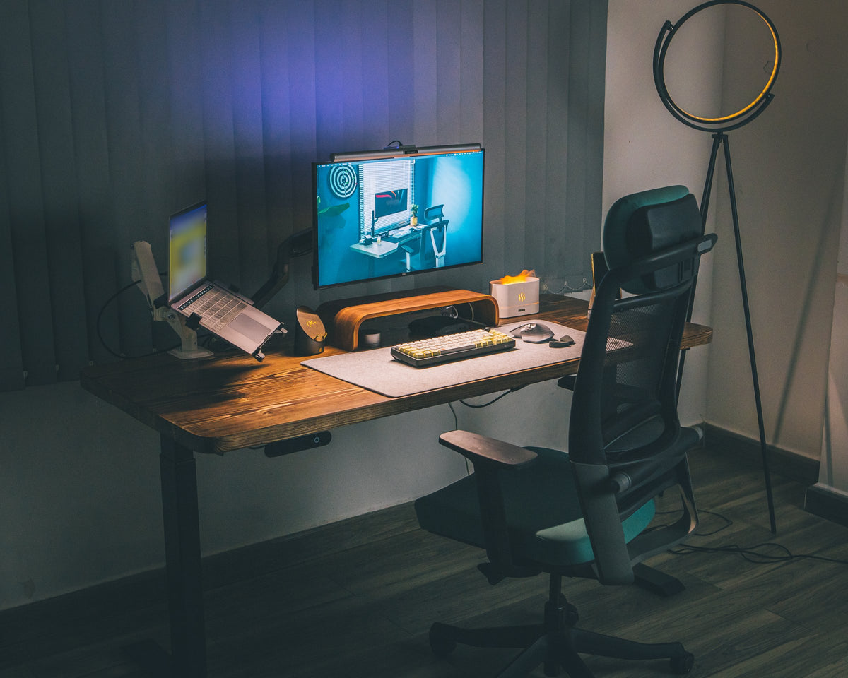 Moody desk setup with a monitor on a wooden riser, laptop on a stand, felt desk mat, keyboard and ring floor lamp