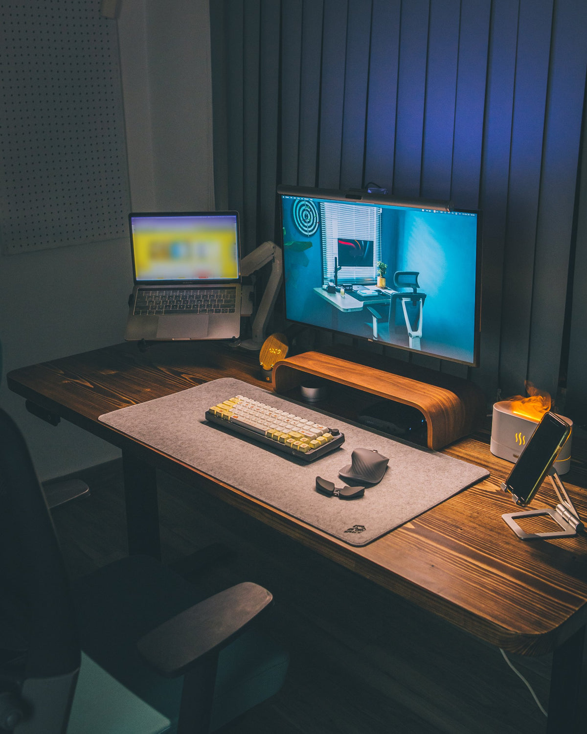 Low-light desk setup with a monitor, laptop on a stand, wooden riser, grey desk mat, keyboard and ergonomic mouse