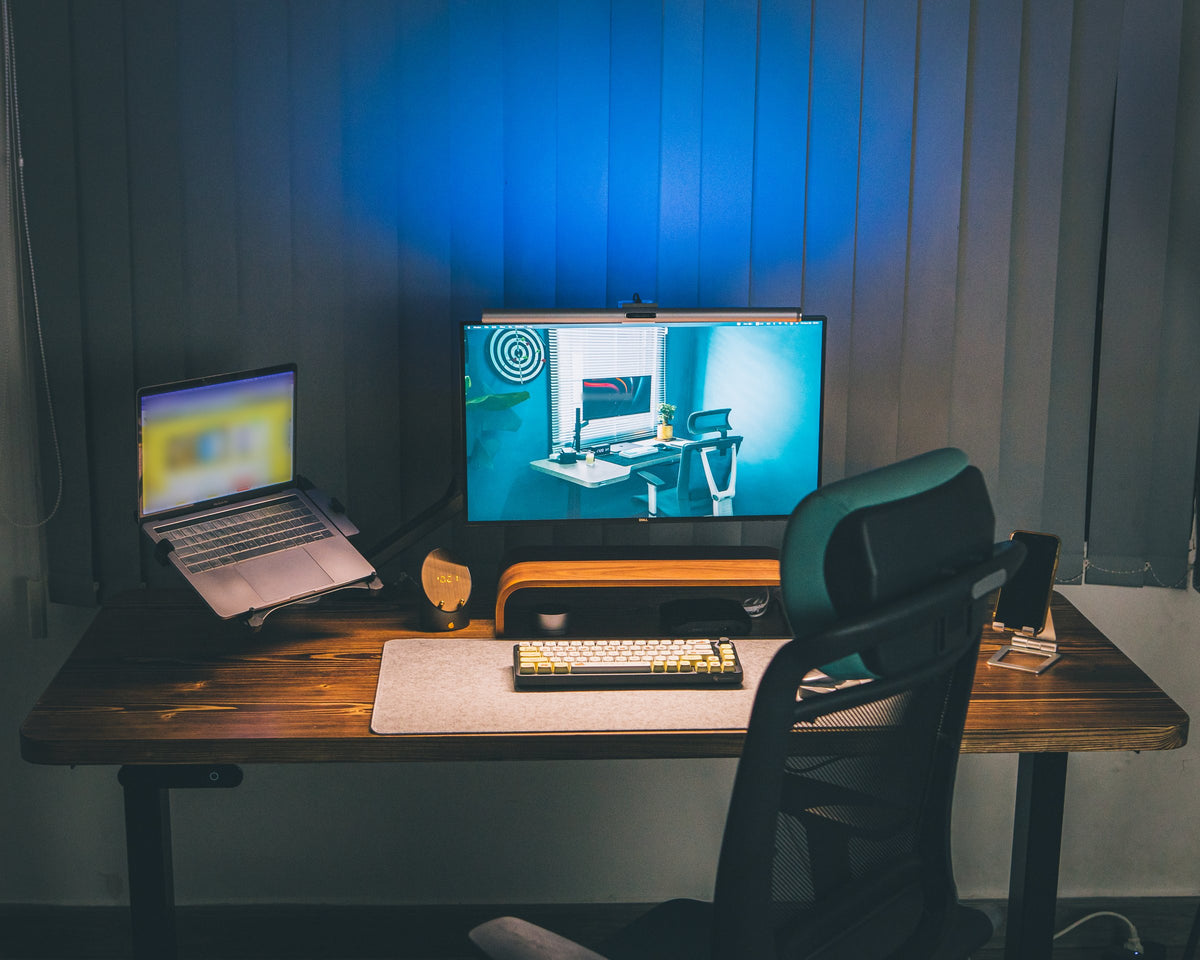 Evening desk setup with a monitor and laptop on a stand, wooden riser, felt desk mat and mechanical keyboard