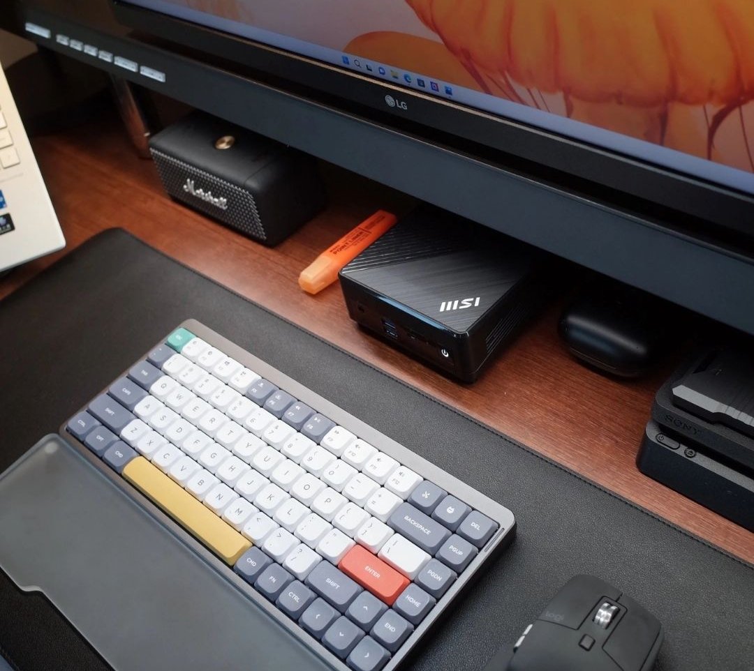 Mechanical keyboard on a black desk mat under a monitor shelf with a mini PC and speaker