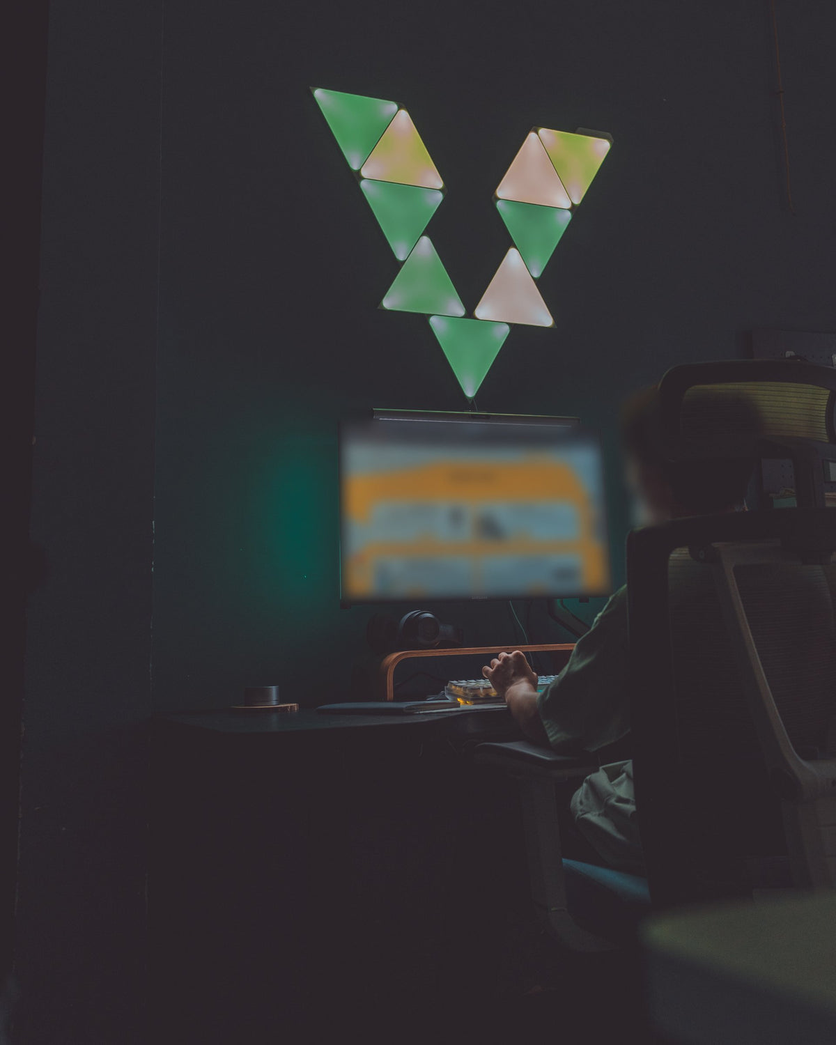 Dark room with a glowing triangle wall light, a monitor on a desk and a person seated at it seen from behind