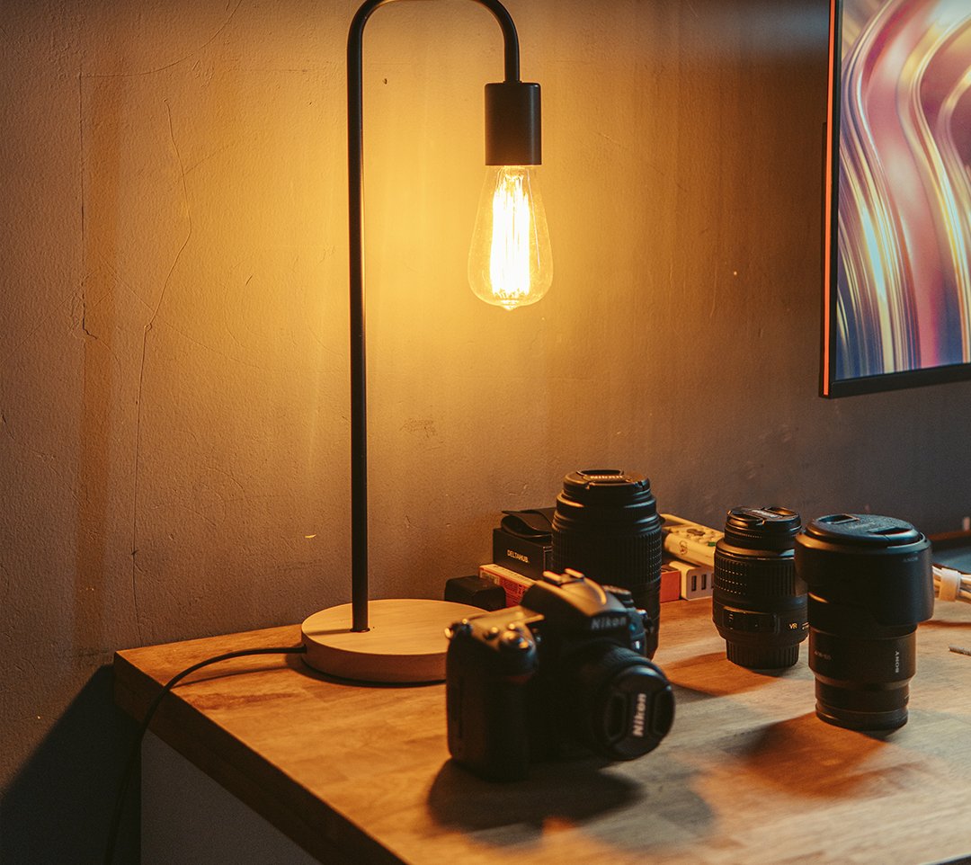 Black table lamp with an Edison bulb next to cameras and lenses on a wood desk