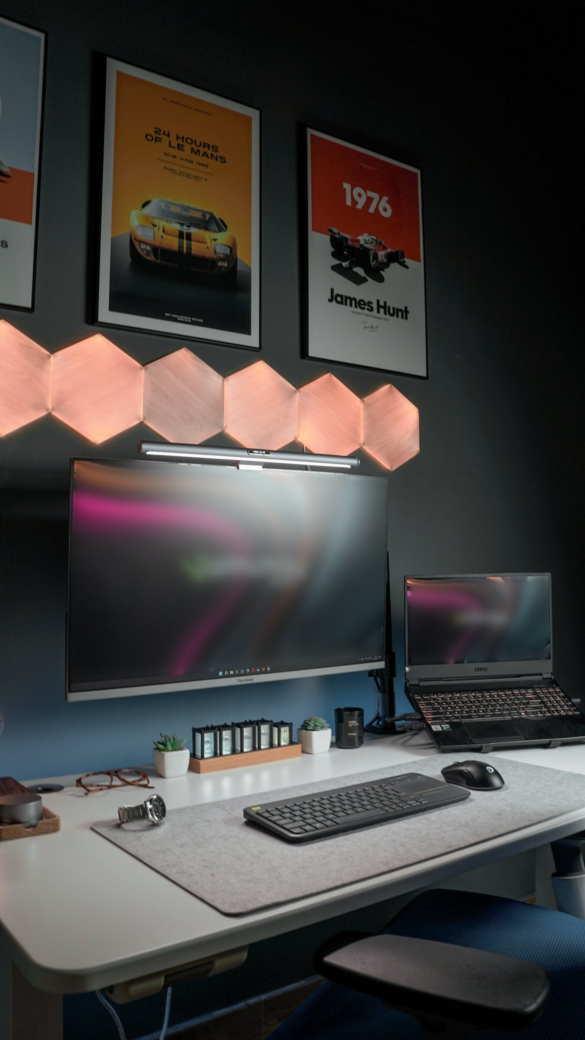 Dark wall with hexagon light panels above a desk with a monitor, laptop on a stand, keyboard and desk mat