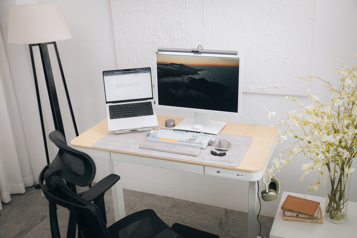 Light oak and white standing desk with a laptop on a stand, a monitor with light bar and a mesh chair