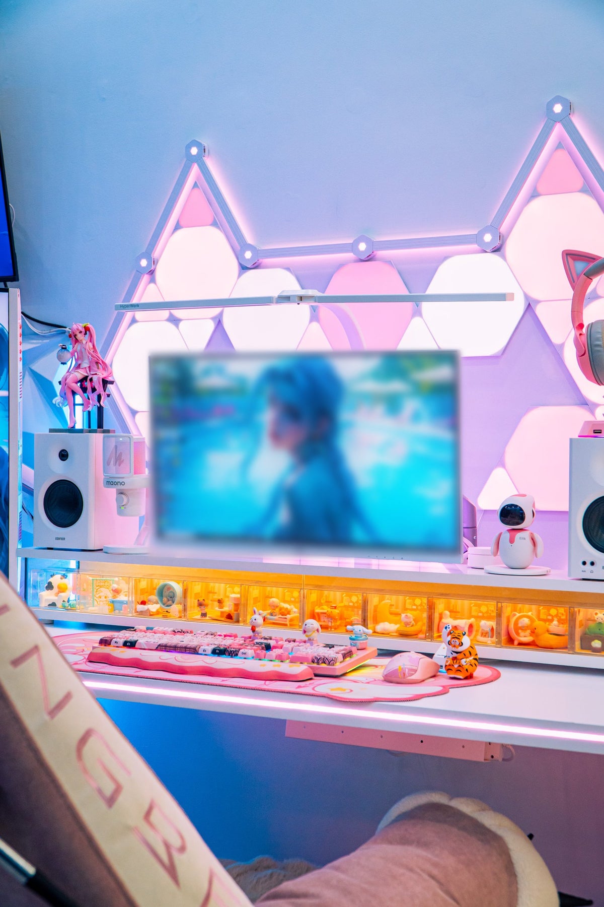 Pink gaming desk under cat-ear hexagon light panels with a display shelf of toy boxes and a plush recliner chair