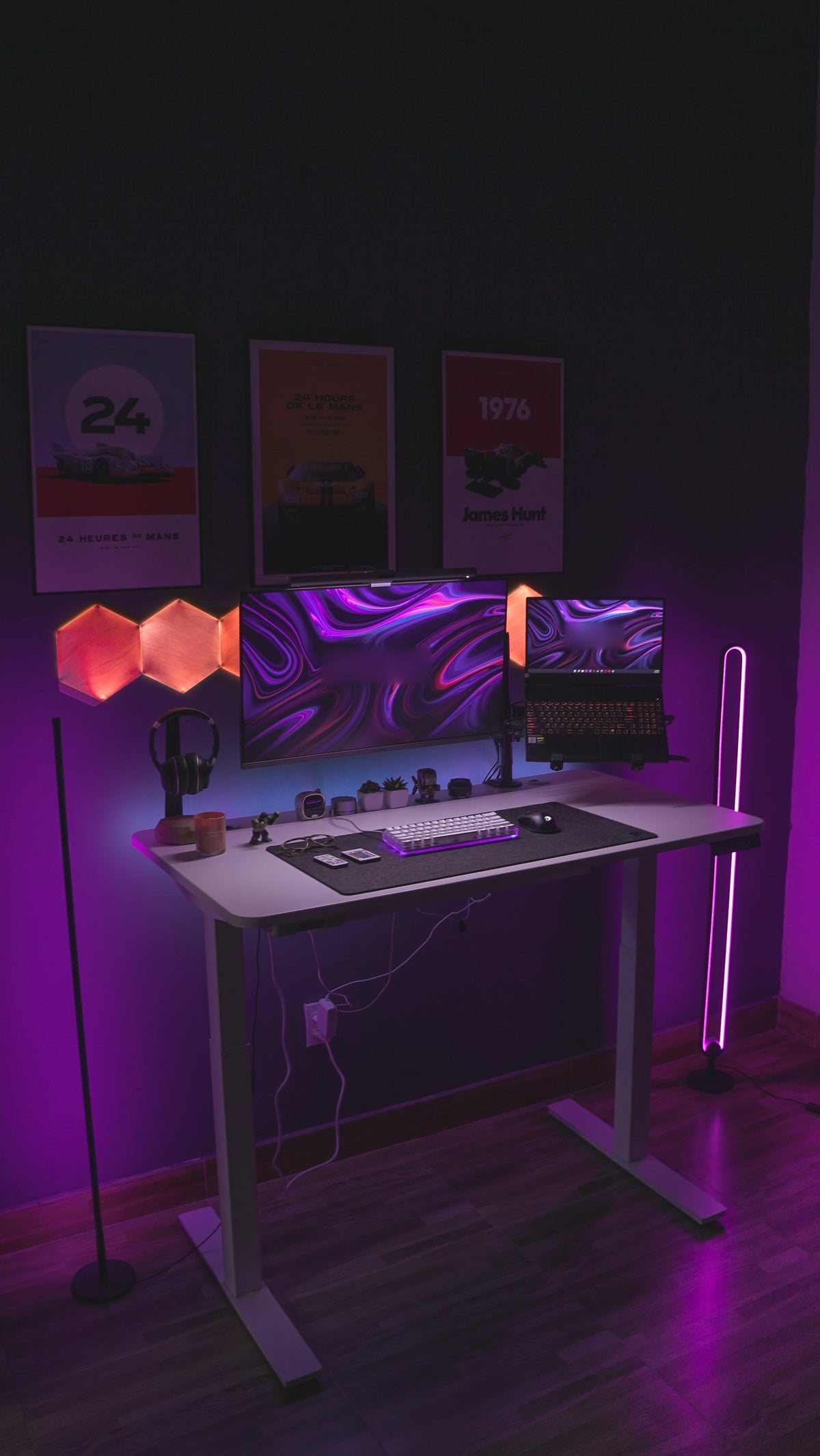 Moody magenta-lit desk setup with monitor, laptop, headphone stand, white keyboard, wall posters and hexagon wall lights