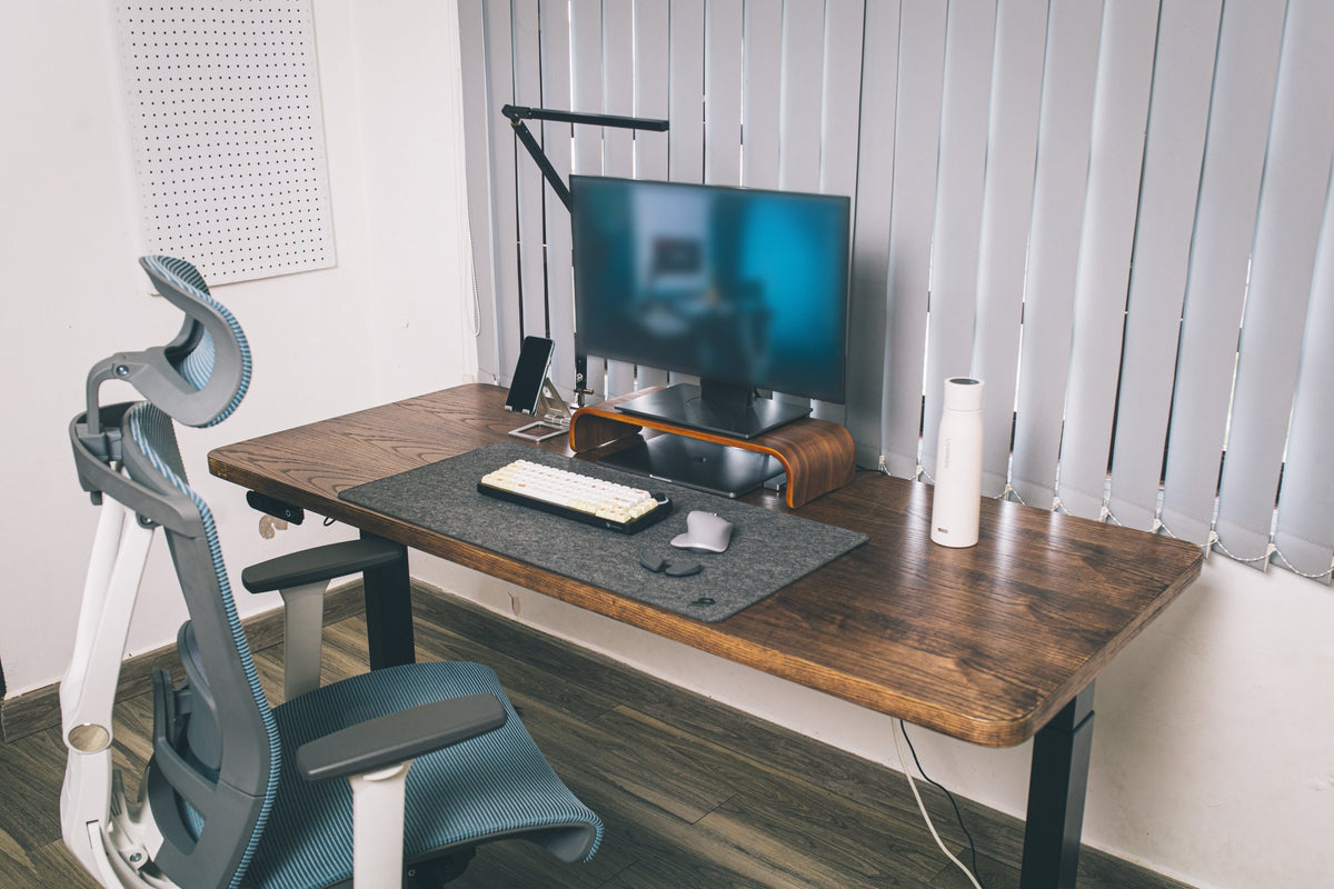 Wooden desk by vertical blinds with monitor on a walnut riser, phone stand, keyboard, mouse, desk mat and ergonomic chair
