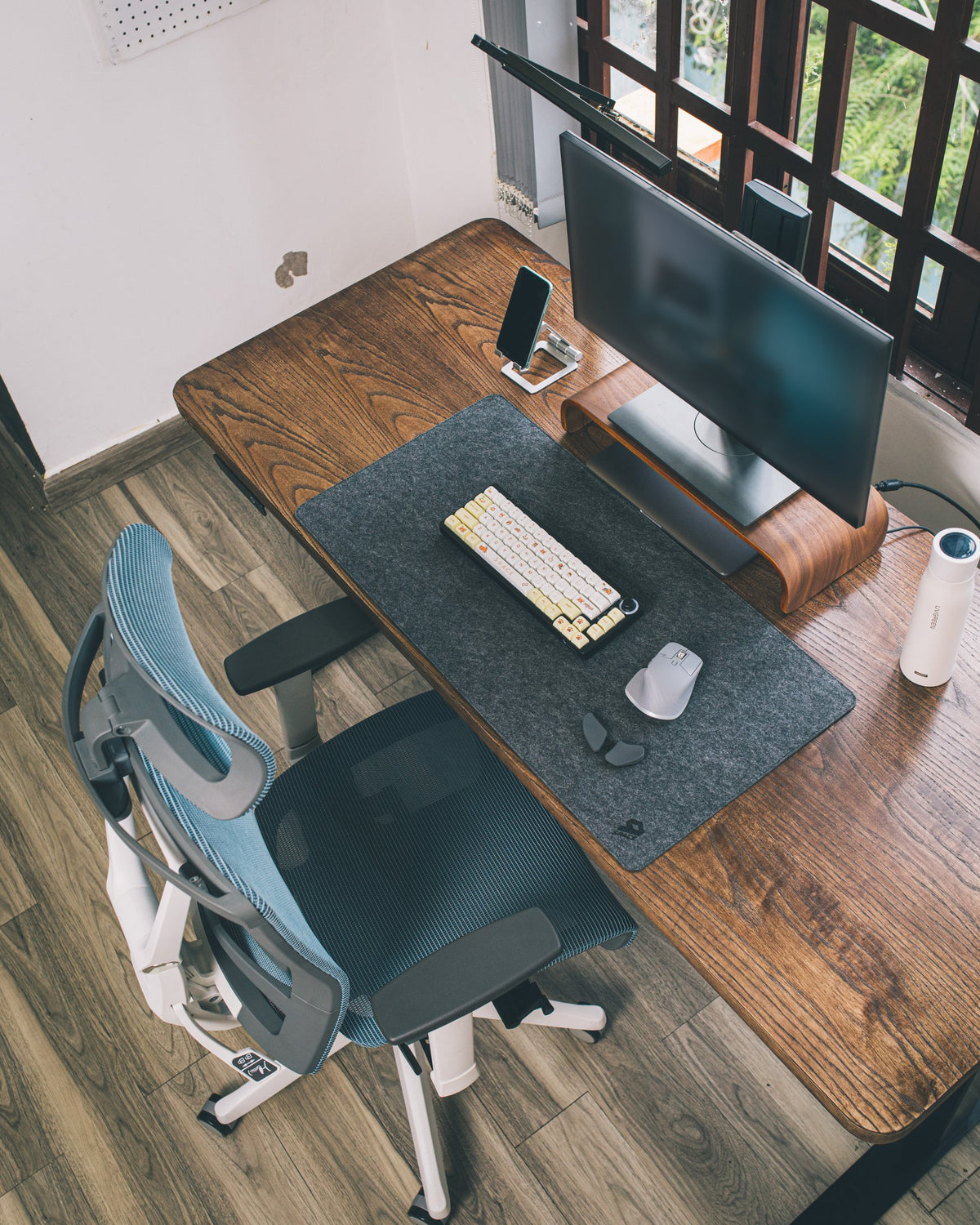 Overhead view of a wooden desk with monitor on a walnut riser, felt desk mat, keycap keyboard, mouse and a mesh ergonomic chair