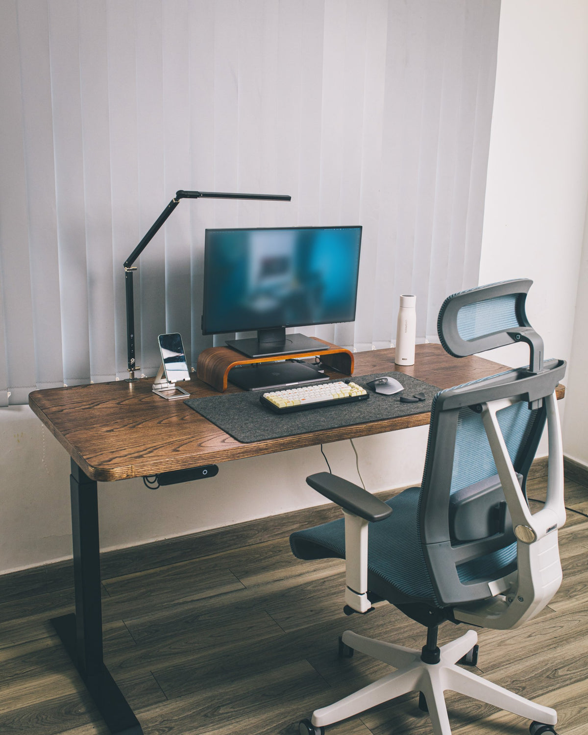 Wooden desk with monitor on a riser, articulated desk lamp, phone stand, white bottle, keyboard and an ergonomic chair