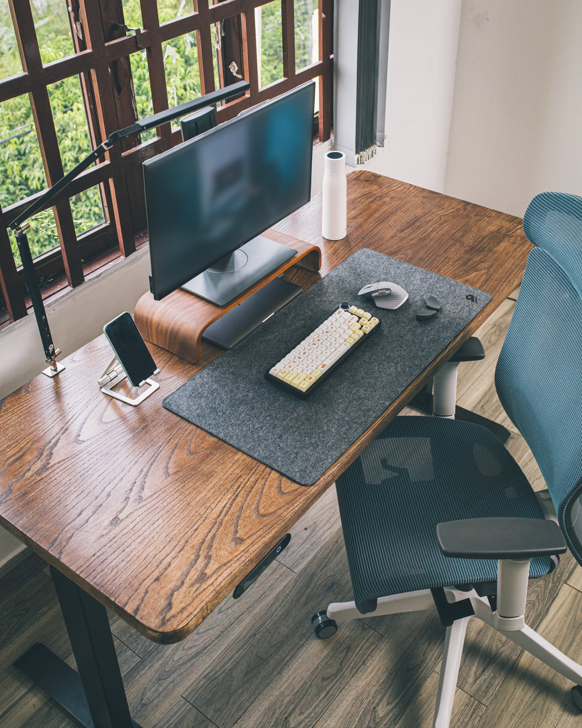 Wooden desk by a window with a monitor on a walnut riser, felt desk mat, mechanical keyboard, mouse and phone stand
