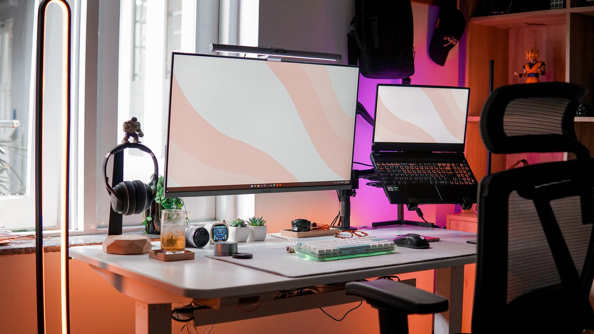 White standing desk with a large monitor and laptop by a sunny window, pink and purple glow