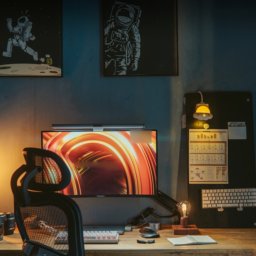 Dimly lit desk with an orange-wallpaper monitor, ergonomic chair, pegboard with a lamp, keyboard and warm bulb on a wooden desktop
