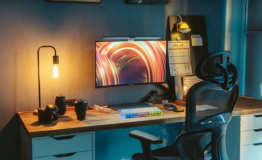 Cozy wooden desk with warm floor lamp, camera and lenses, monitor with orange wallpaper, keyboard and ergonomic chair