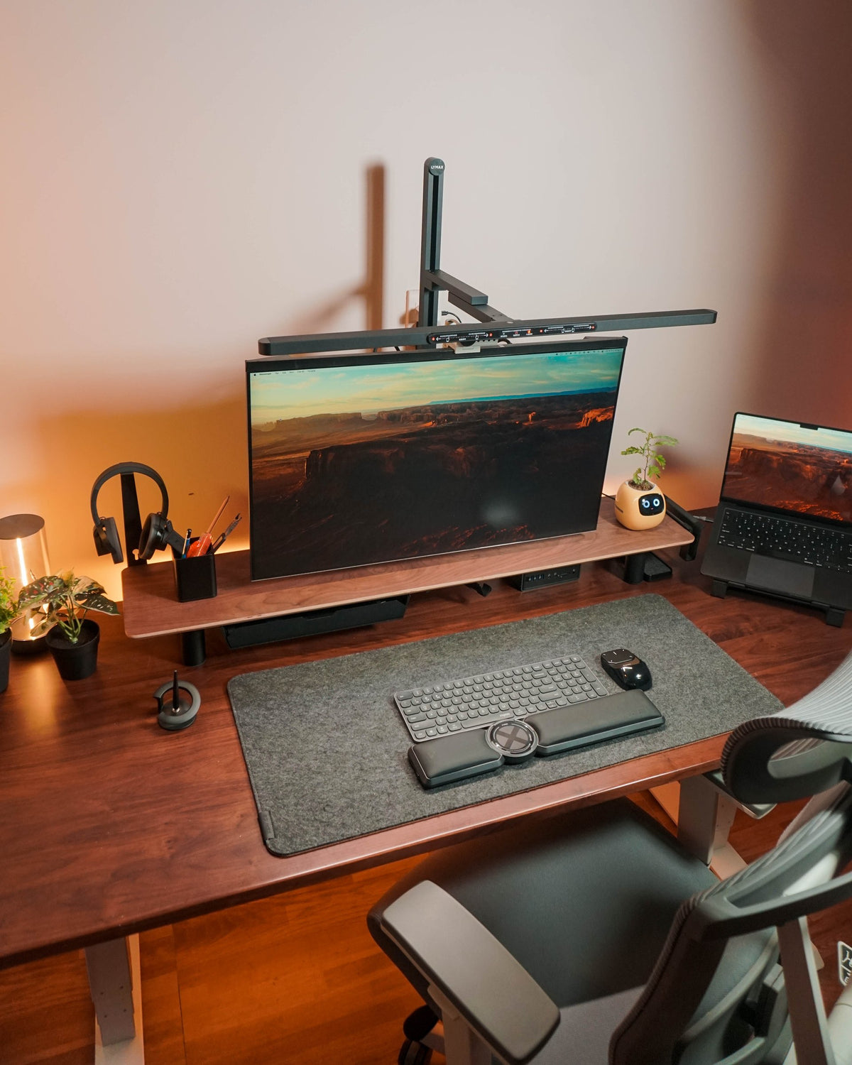Walnut standing desk with a grey felt mat, keyboard with wrist rest and a large monitor under an arm lamp