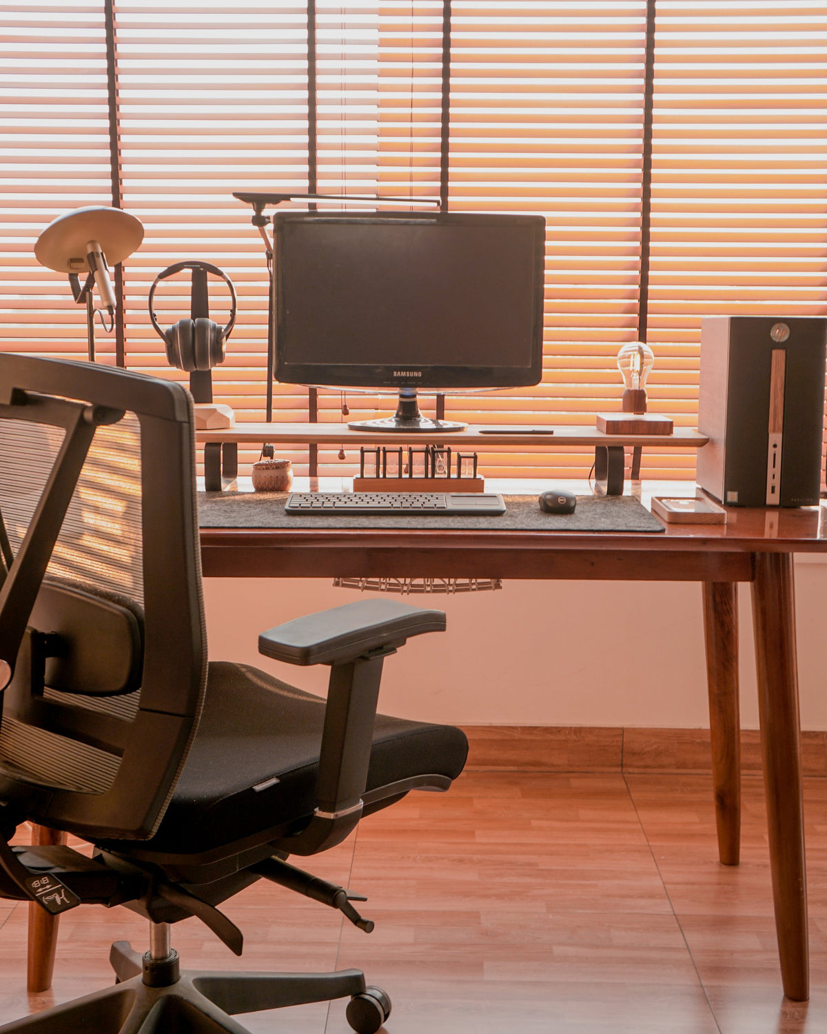 Wood desk with a monitor riser shelf and mesh chair in front of sunlit blinds