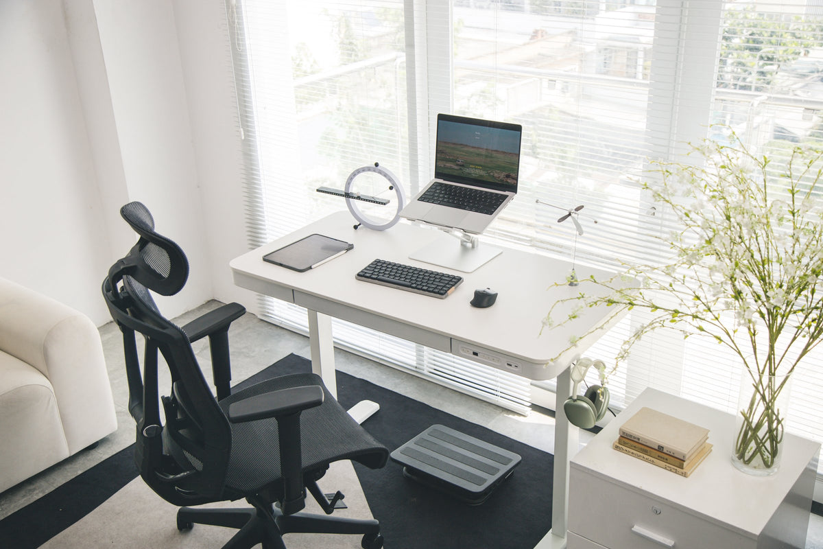 White minimalist standing desk by the window with a laptop on a stand, ring desk clock and black mesh chair