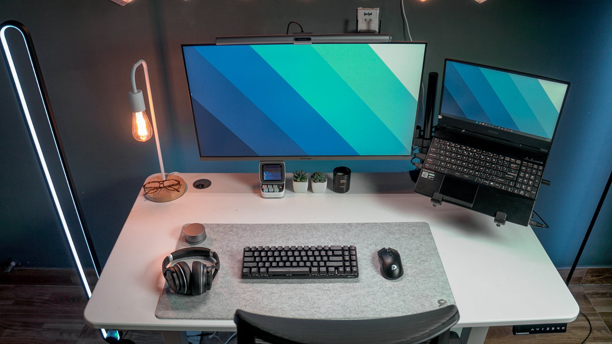 White desk with a large monitor, laptop on an arm, Edison lamp and headphones on a felt mat