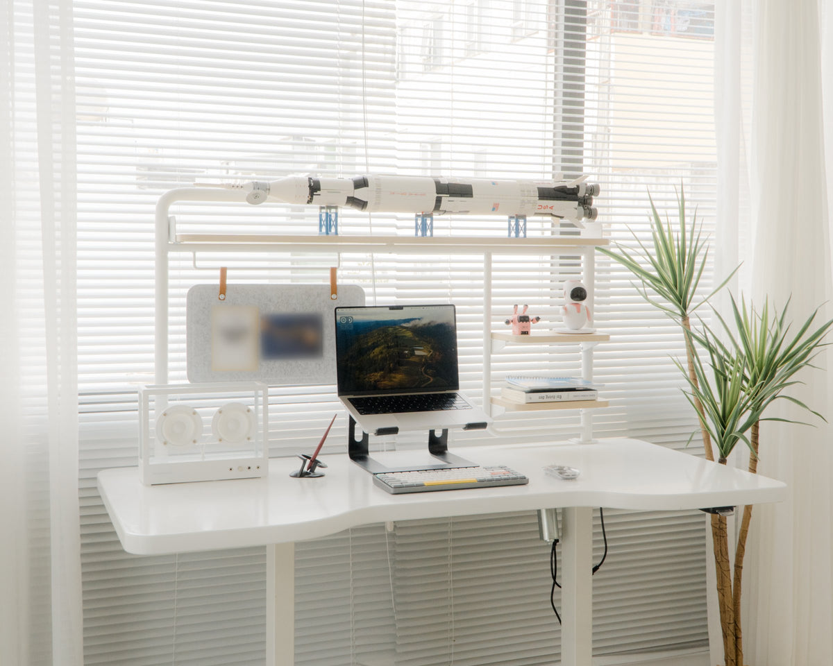 White standing desk by a blinds window with laptop on a stand, speaker, felt pinboard and a model rocket on the shelf