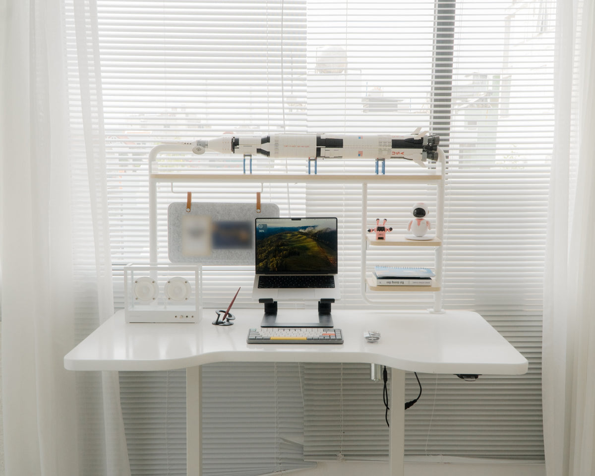 Bright minimalist white desk with laptop on riser, keyboard, speaker, felt pinboard and small robot figures on a shelf