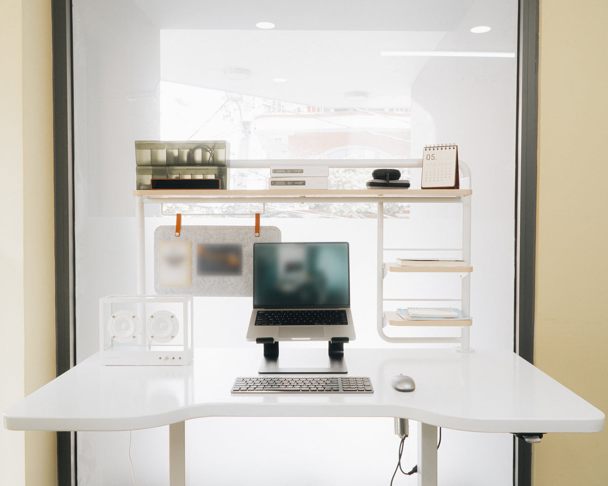 White desk with laptop on a stand, wireless keyboard, mouse, speaker, desk calendar and open shelves against a bright window