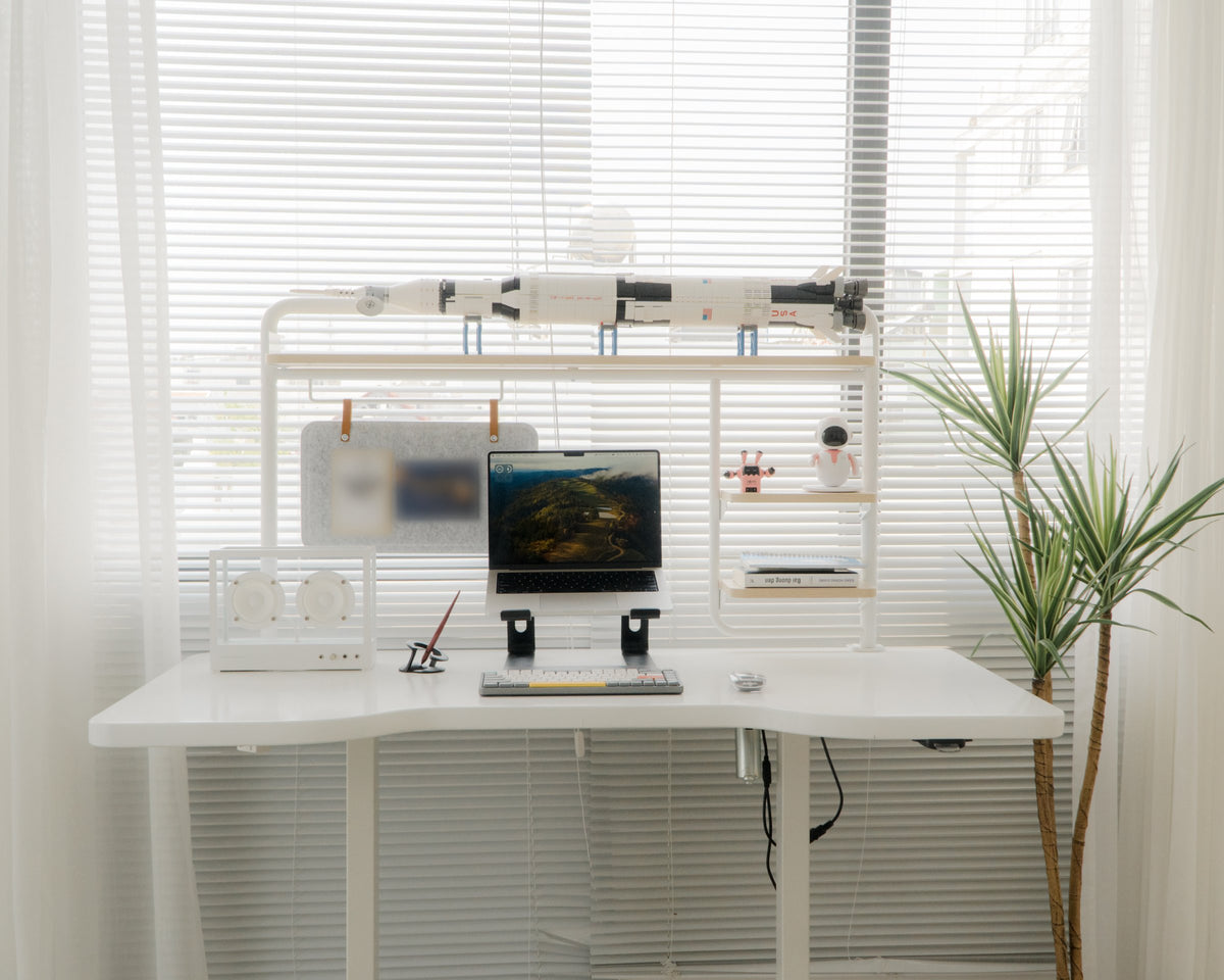 White desk setup in front of a window with laptop, mechanical keyboard, desktop speaker and rocket model on an overhead shelf