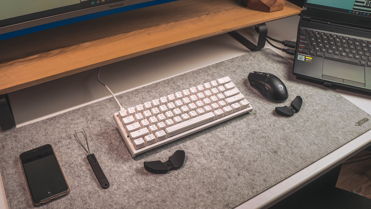 White mechanical keyboard, black mouse and phone on a gray felt desk mat under a wood shelf