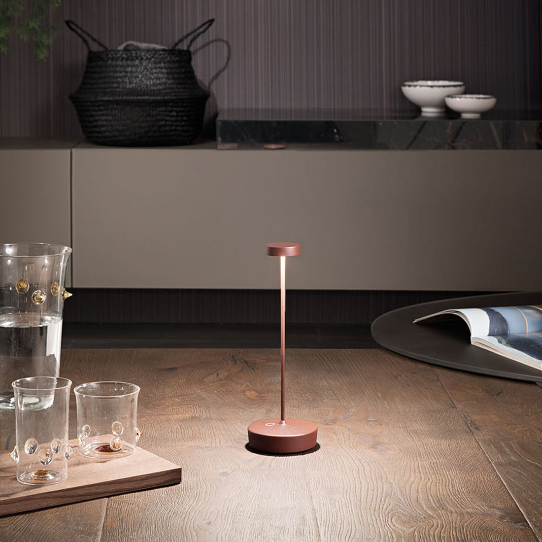 A copper-toned Swap Pro lamp lit on a wooden floor beside glass cups and a magazine