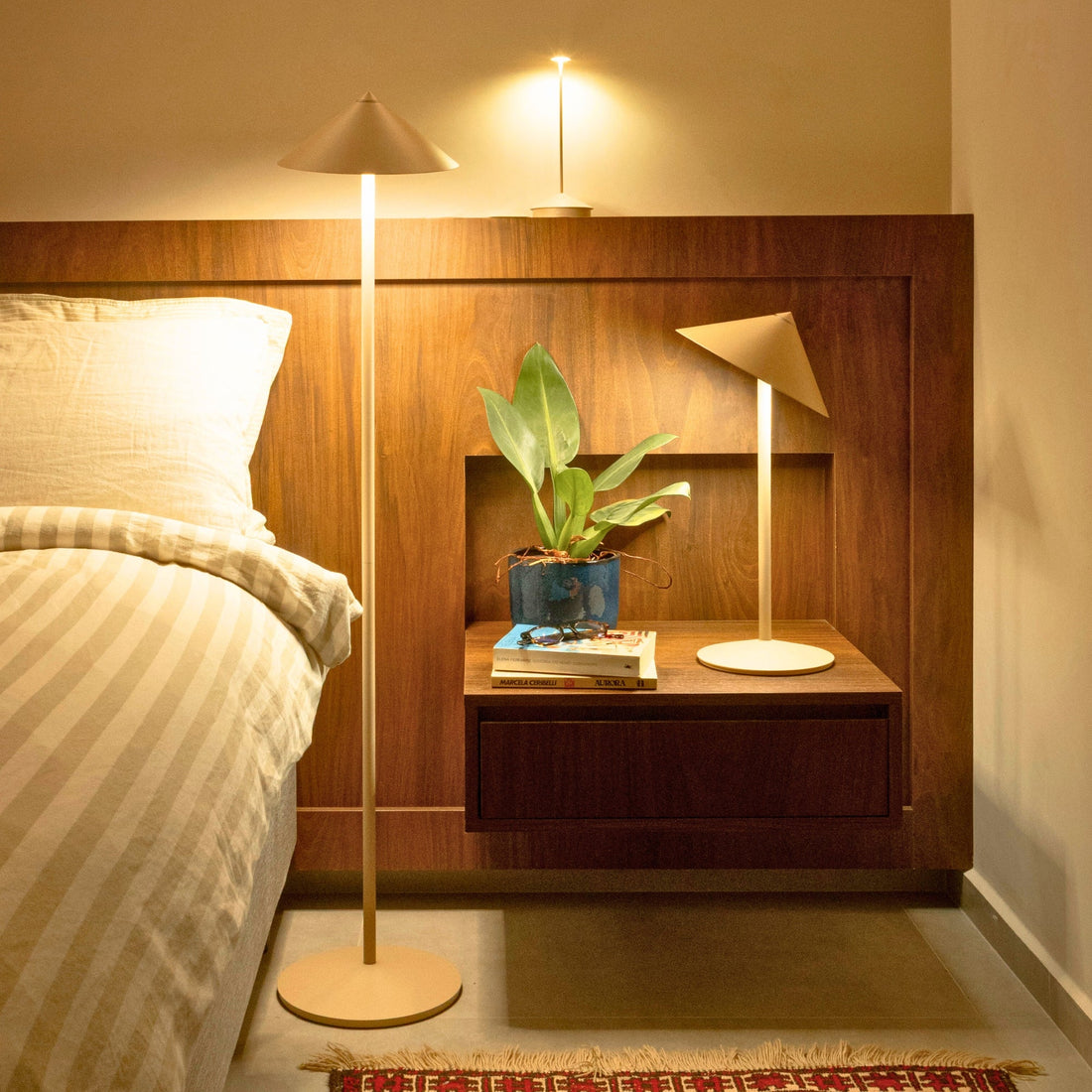A Pina lamp glowing warm on a bedside shelf in a bedroom, a taller floor version beside the bed