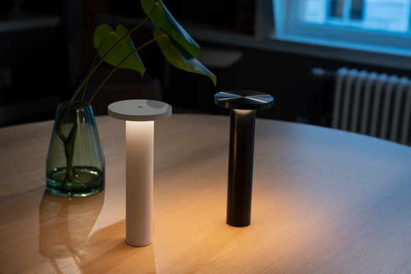 A white and a black Luci lamp lit beside a glass vase with a leaf on a wooden table