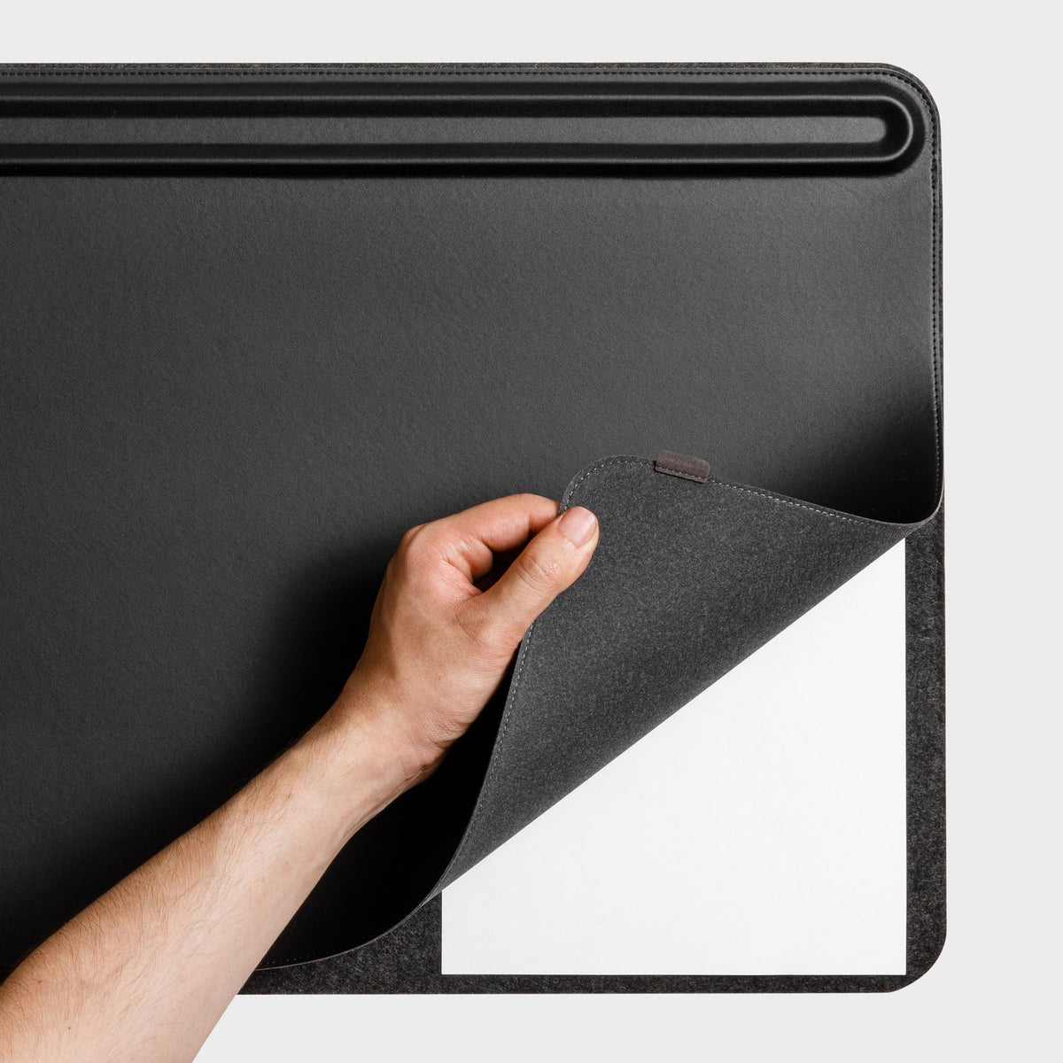 Hand lifting the corner of the black desk mat to show a white sheet of paper underneath