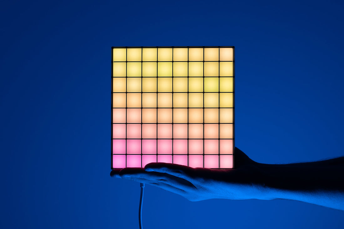 A hand holding one lit square LED panel with a warm yellow to pink gradient against a blue backdrop