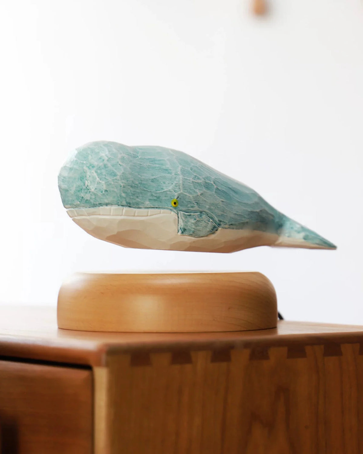 A carved whale with a sea-blue back and a white belly floats just above a round pale wood base on a wooden cabinet.