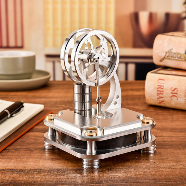 Silver metal Stirling engine with two flywheels on a wooden desk beside books and a cup