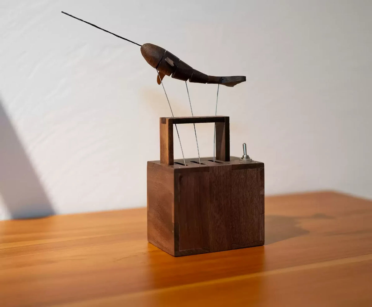 The narwhal automaton on a wooden table against a white wall, its horn pointing up and to the left.