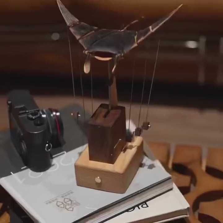 Side view of the manta ray automaton, dark wings above the tower and a pale wood base, a camera and a book behind.